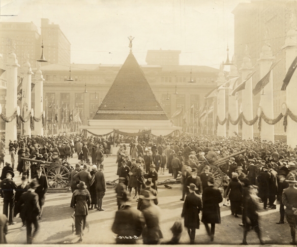 "View of the employees of the New York Central Railroad, assembled in Victory Way, showing the pyramid of captured German helmets, with Grand Central Terminal in the background."